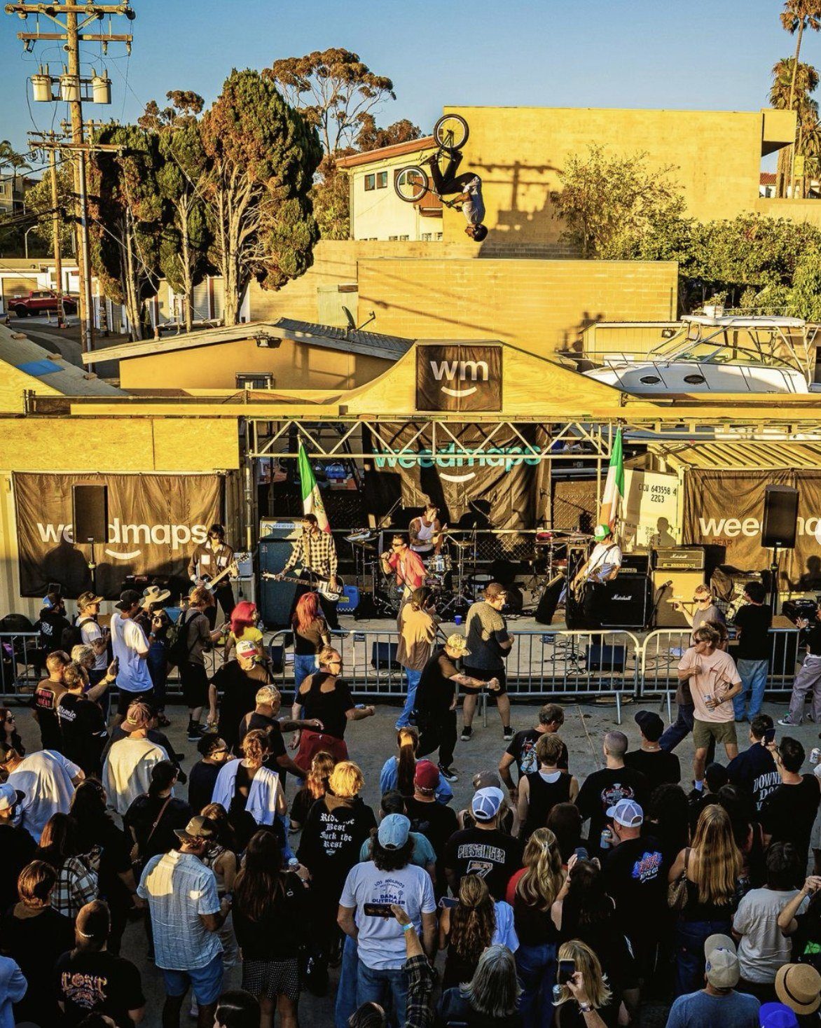 A BMX rider inverted mid-air above the Weedmaps stage at AMAC (Art Music Athlete Collective) at golden hour, band playing to a packed crowd.